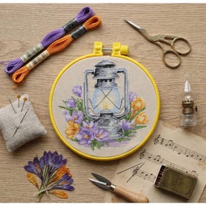 Può includere: Un telaio da ricamo a punto croce con un design di lanterna vintage circondato da fiori viola e gialli. La scena comprende filo da ricamo, forbici, un cuscinetto per spilli, una piccola lampada a olio e spartiti, il tutto disposto su una superficie di legno.