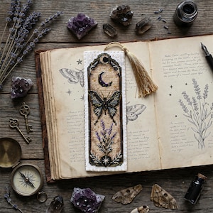 Peut inclure: Un marque-page rectangulaire en point de croix sur tissu beige représentant un papillon noir, un croissant de lune violet et des brins de lavande. Le marque-page est bordé d'un cadre noir avec des détails dorés et un pompon doré fixé au sommet.
