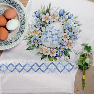 May include: White embroidered kitchen towel with a blue and white floral design featuring a large egg in the centre. The towel has a blue and white diamond pattern border and fringe.