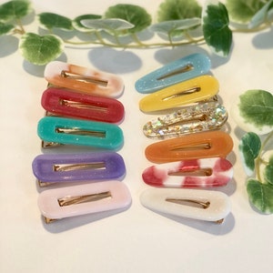 Peut inclure: Un ensemble de dix pinces à cheveux colorées. Les pinces sont en plastique et présentent une variété de motifs, notamment des paillettes, du tie-dye et des couleurs unies. Les pinces sont toutes en forme de rectangle aux bords arrondis.