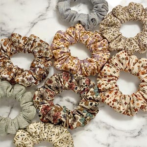 May include: A collection of scrunchies in various floral patterns and solid colors. The scrunchies feature shades of brown, pink, purple, and green, with a gray ribbed scrunchie at the top. The scrunchies are made of fabric and are designed for hair.