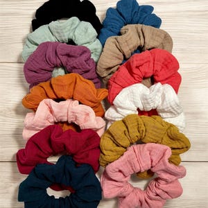 May include: A stack of 12 colorful scrunchies made of soft, textured fabric. The scrunchies are in various colors, including black, teal, light green, purple, orange, white, pink, red, dark blue, and light pink.