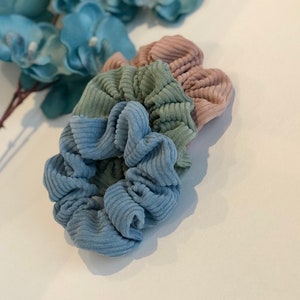 Puede incluir: Tres scrunchies de tela de pana azul, verde y rosa. Los scrunchies est&aacute;n apilados uno encima del otro.