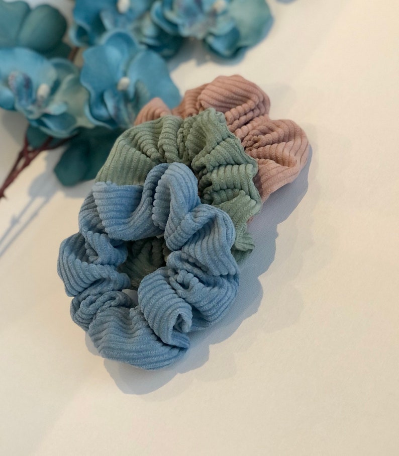 Puede incluir: Tres scrunchies de tela de pana azul, verde y rosa. Los scrunchies est&aacute;n apilados uno encima del otro.