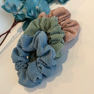 Puede incluir: Tres scrunchies de tela de pana azul, verde y rosa. Los scrunchies est&aacute;n apilados uno encima del otro.
