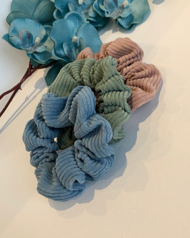 Puede incluir: Tres scrunchies de tela de pana azul, verde y rosa. Los scrunchies est&aacute;n apilados uno encima del otro.