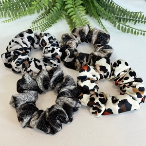 May include: Four scrunchies in different animal print patterns. One is black and white, one is brown and white, one is black and gray, and one is white with brown and orange spots.