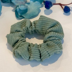 Puede incluir: Un scrunchie de color verde salvia hecho de tela de pana. El scrunchie est&aacute; recogido y fruncido, creando un aspecto suave y voluminoso.