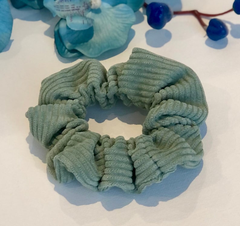 Puede incluir: Un scrunchie de color verde salvia hecho de tela de pana. El scrunchie est&aacute; recogido y fruncido, creando un aspecto suave y voluminoso.