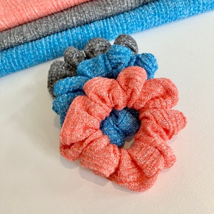 May include: Three scrunchies in coral, blue, and gray. The scrunchies are made of a soft, textured fabric.