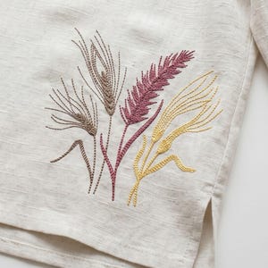 May include: Close-up of a beige linen fabric with an embroidered design of wheat stalks. The embroidery features shades of brown, pink, and yellow, creating a natural and textured appearance. The fabric has a subtle, woven pattern.