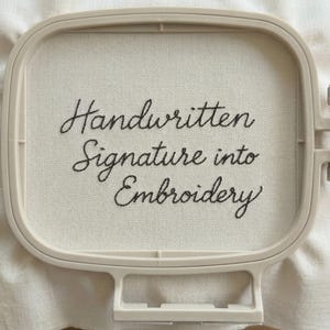 May include: A cream-colored embroidery hoop holding white fabric with the words "Handwritten Signature into Embroidery" stitched in black thread. The hoop is a light beige color.