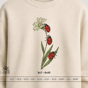 May include: A beige crewneck sweatshirt featuring an embroidered design of a green stem with a white flower and three red ladybugs with black spots. The text "AMER EMBROIDERY STUDIO", "5x7 - 6x10", and file formats "DST - PES - VP3 - VIP - HUS - EXP - XXX - EMP - JEF - BMP" are printed below the design.