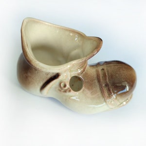 May include: A ceramic shoe-shaped planter in shades of brown and beige. The planter has a rounded toe, a high collar, and a small hole on the side. The interior is a lighter beige colour.