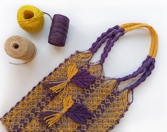 Può includere: Una borsa a mano in crochet viola e gialla con un manico annodato. La borsa è realizzata con diversi punti a crochet e ha un design unico. La borsa è su una superficie bianca con tre gomitoli di lana.