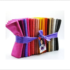 Puede incluir: Una pila de 20 cuadrados de tela coloridos atados con una cinta morada. Los cuadrados de tela son en tonos de rosa, rojo, naranja, amarillo y gris. Una etiqueta con el texto "Miss P Fabrics" está unida a la cinta.