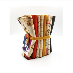 Op de afbeelding: Een bundel van acht stofvierkanten met verschillende patronen, waaronder kippen, tekst en geometrische ontwerpen. De stoffen zijn in tinten bruin, geel, oranje en wit. De bundel is vastgebonden met een geel lint en heeft een klein label met de tekst "Miss P Fabrics".