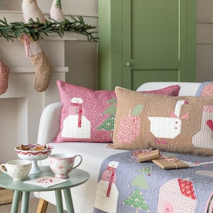 May include: A white couch with a gray and pink quilt and three pillows. The pillows are decorated with a snowman, a goose, and a Christmas tree. The couch is in front of a fireplace with stockings hanging from the mantle. There is a green cabinet in the background.