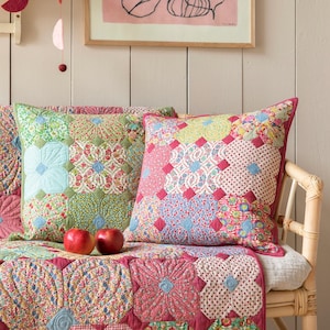 Könnte beinhalten: Gemütliche Inneneinrichtung mit einer floralen Steppdecke und zwei dekorativen Kissen mit bunten Patchworkmustern. Ein gerahmtes Kunstwerk mit rosa Hintergrund hängt darüber. Zwei rote Äpfel liegen auf der Steppdecke.