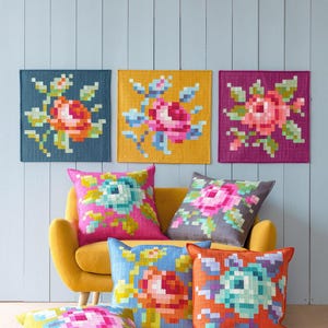 Może przedstawiać: Kolekcja kwadratowych poduszek dekoracyjnych i dekoracji ściennych z pikselowanymi wzorami kwiatowymi. Paleta kolorów obejmuje odcienie różu, niebieskiego, pomarańczowego i zielonego. Poduszki ułożone są na żółtej sofie, a dekoracje ścienne zawieszone na jasnoniebieskiej ścianie.