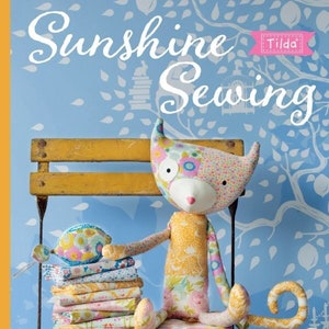 Puede incluir: Un libro titulado "Sunshine Sewing" de Tone Finnanger con un logotipo de Tilda. La portada presenta un gato de tela colorido sentado en un banco de madera con una pila de recortes de tela.