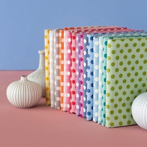 Op de afbeelding: Een stapel van 14 stofvierkanten met een wit stippenpatroon op verschillende gekleurde achtergronden, waaronder geel, oranje, rood, roze, paars, blauw en groen. De stofvierkanten zijn in een regenboogpatroon gerangschikt en liggen op een roze oppervlak.