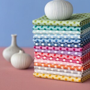 Op de afbeelding: Een stapel kleurrijke stofvierkanten met witte stippen, gerangschikt in een regenboogpatroon. Een witte keramische vaas met een textuur zit bovenop de stapel.