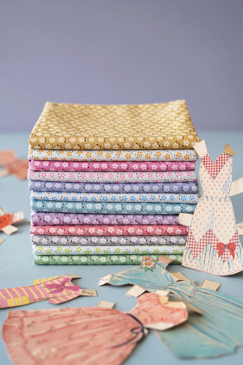 May include: A stack of 12 fabric squares with different floral patterns in shades of yellow, pink, purple, blue, and green. The fabric squares are stacked on a light blue surface. There are paper dolls with dresses on the surface.