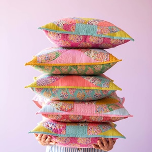 Puede incluir: Una pila de cinco almohadas de patchwork coloridas con patrones florales. Las almohadas están dispuestas en forma de pirámide y se sostienen frente a una persona que lleva una camisa a rayas.