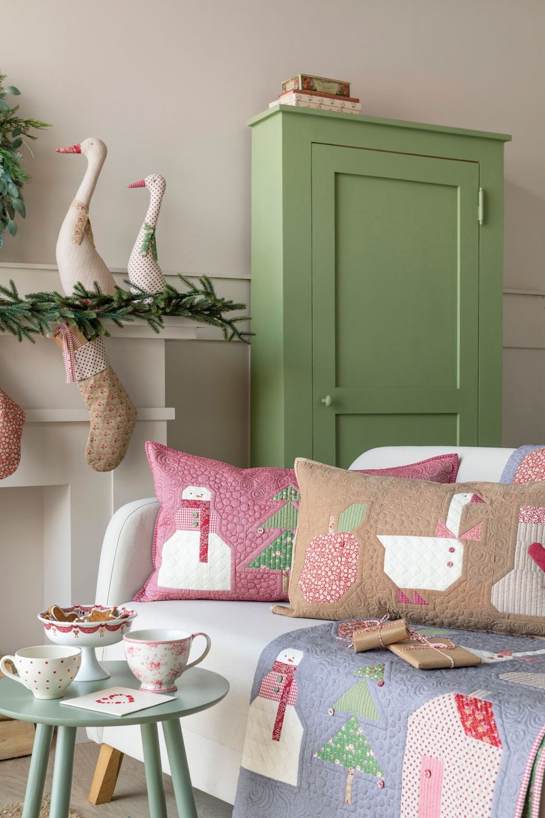 May include: A white couch with a gray and pink quilt and three pillows. The pillows are decorated with a snowman, a goose, and a Christmas tree. The couch is in front of a fireplace with stockings hanging from the mantle. There is a green cabinet in the background.