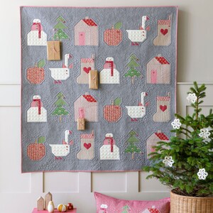 May include: A gray quilted wall hanging with a Christmas theme features a variety of festive designs, including houses, snowmen, geese, apples, and Christmas trees. The wall hanging is decorated with pink and red accents and is surrounded by a white border. A small Christmas tree with white ornaments is in the foreground.