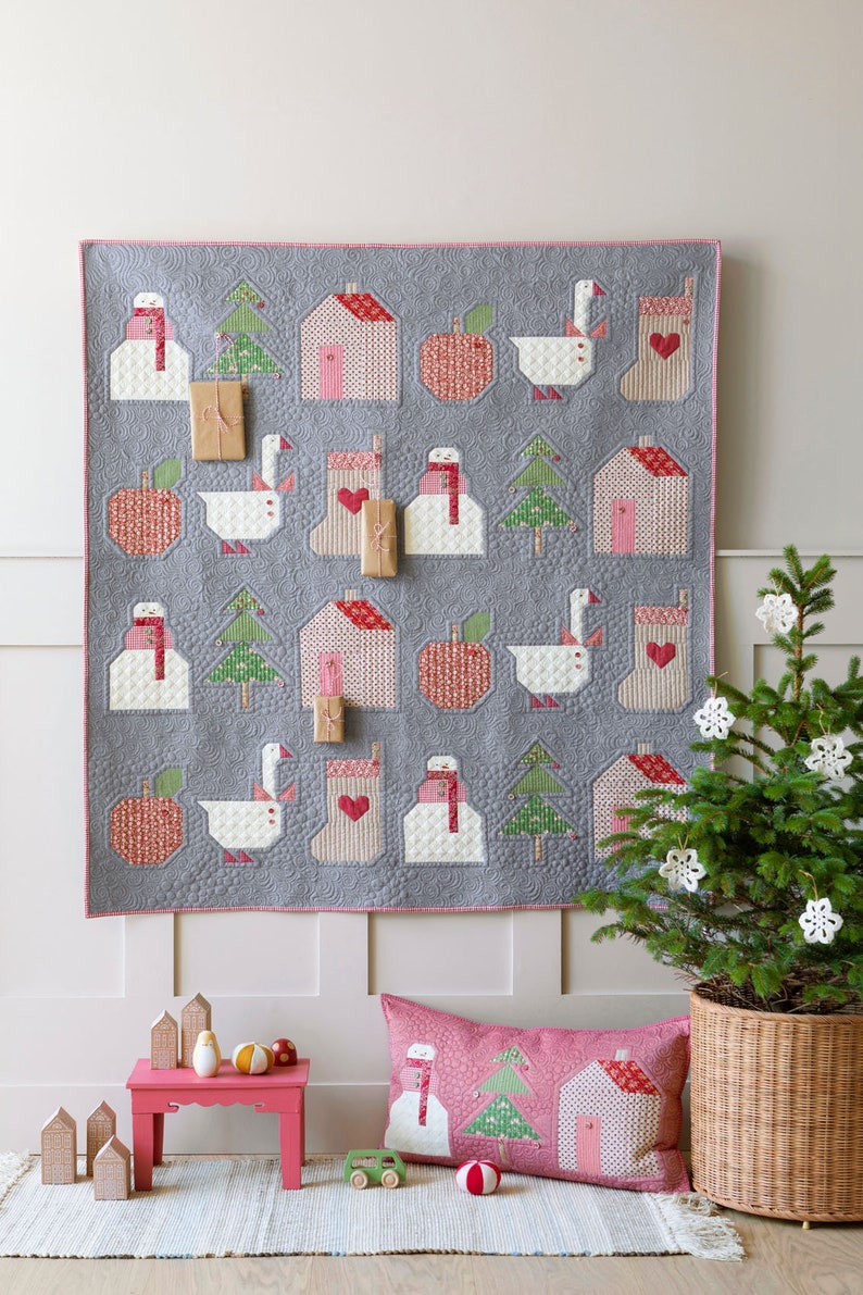 May include: A gray quilted wall hanging with a Christmas theme features a variety of festive designs, including houses, snowmen, geese, apples, and Christmas trees. The wall hanging is decorated with pink and red accents and is surrounded by a white border. A small Christmas tree with white ornaments is in the foreground.