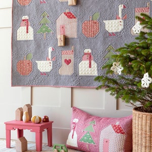 May include: A pink and white patchwork pillow with a snowman, a Christmas tree, and a house design. The pillow is on a wooden floor with a book and a red and white ball.