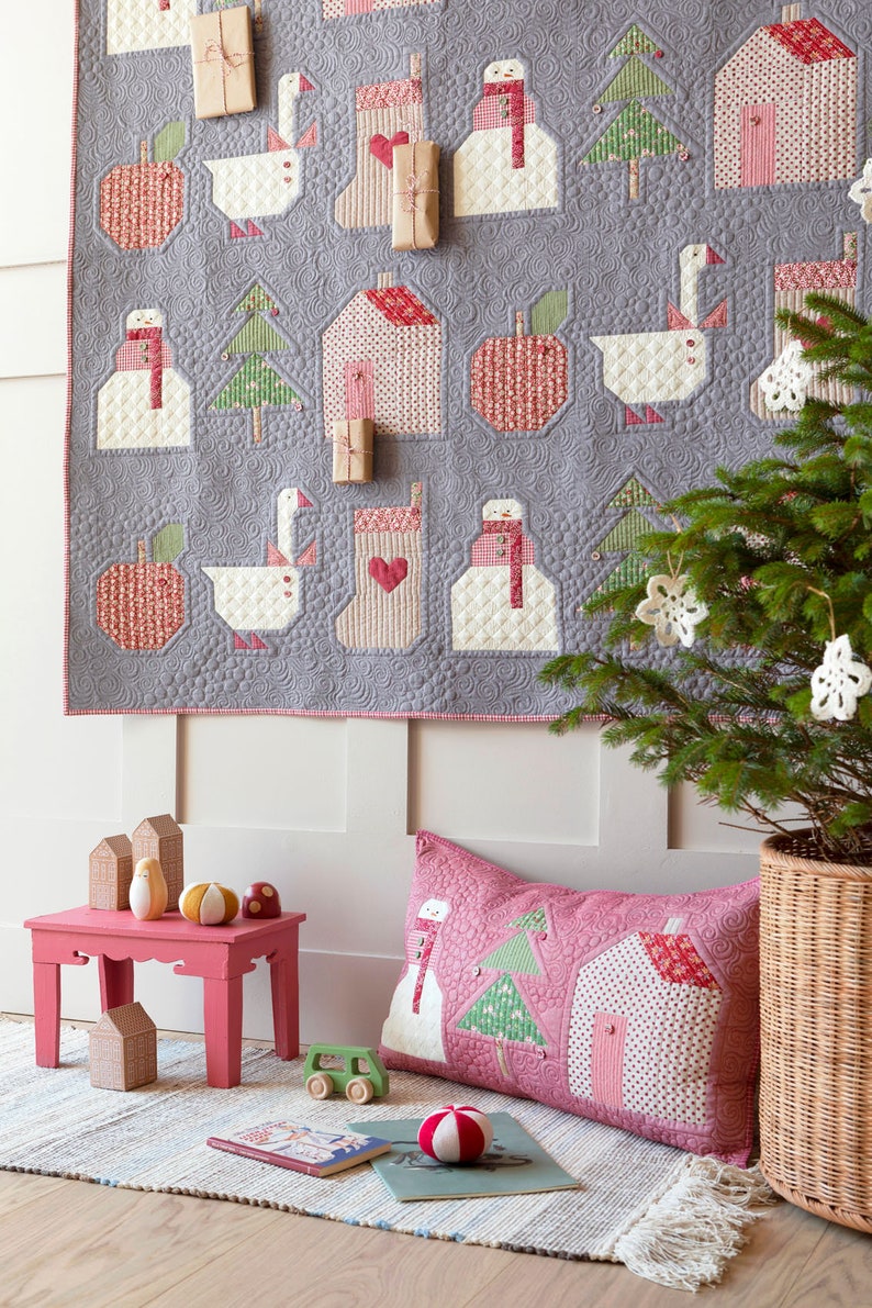 May include: A pink and white patchwork pillow with a snowman, a Christmas tree, and a house design. The pillow is on a wooden floor with a book and a red and white ball.