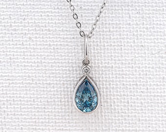 May include: A silver necklace with a teardrop-shaped, light blue pendant. The pendant is accented with a small, round, clear stone above the blue stone. The necklace is displayed on a white textured surface.