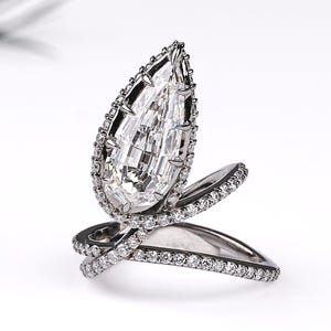 Puede incluir: Un anillo de metal oscuro con una gran piedra central en forma de pera rodeada por un halo de pequeñas piedras claras. La banda es abierta, estilo bypass, e incrustada con una hilera de pequeñas piedras claras a lo largo del cuerpo.