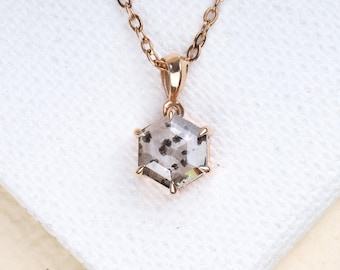 May include: A rose gold necklace featuring a hexagonal pendant with a clear, speckled gemstone. The pendant is suspended from a delicate chain, resting on a white fabric surface. The necklace is a piece of jewelry.