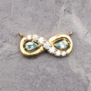 May include: A gold-toned infinity symbol pendant featuring a row of clear round-cut stones along the center and two pear-shaped light green stones set within the loops. The piece includes two small metal loops at the top for attachment.