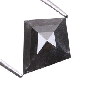Könnte beinhalten: Ein quadratisch geschliffener, schwarzer Diamant, der von einer silbernen Pinzette gehalten wird. Der Diamant hat eine facettierte Oberfläche mit einem reflektierenden, geometrischen Muster in der Mitte. Der Edelstein ist dunkelgrau.