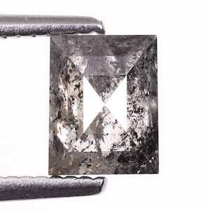Puede incluir: Un diamante de talla rectangular, con aspecto de sal y pimienta, sujeto con pinzas metálicas. El diamante presenta inclusiones grises y negras, con un diseño geométrico blanco en el centro. Sobre un fondo blanco.