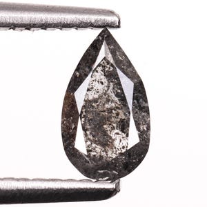 以下が含まれることがあります： 銀色のピンセットで保持された、洋梨型のソルトアンドペッパーダイヤモンド。 ダイヤモンドは、内部のインクルージョンと光を捉えるファセットを持つ、濃いグレー色をしています。 宝石は白い背景にあります。