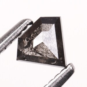 Peut inclure: Un diamant noir taillé en forme de bouclier, maintenu par une pince métallique. Le diamant présente des facettes grises foncées et claires, reflétant la lumière. Le bijou est posé sur un fond blanc.