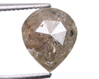Puede incluir: Un diamante en bruto en forma de pera, sujeto por unas pinzas metálicas. El diamante tiene un tono marrón claro con un diseño geométrico facetado en la parte superior. La superficie de la piedra tiene un aspecto texturizado, con inclusiones visibles.