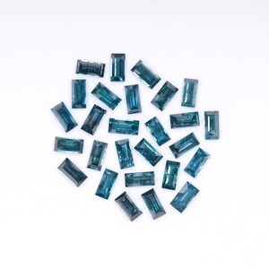 Op de afbeelding: Een verzameling van 20 blauwgroene edelstenen, gesneden in rechthoekige vormen, gerangschikt in een losse cirkel op een witte achtergrond.