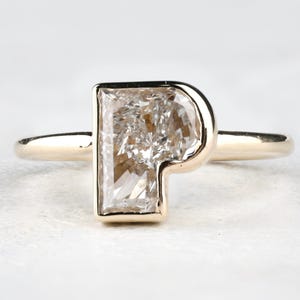 Pode incluir: Um anel de ouro com um grande diamante retangular. O diamante está engastado em uma moldura de bisel, com a banda de ouro formando o haste do anel. O diamante tem uma estrutura interna irregular, dando-lhe uma aparência única.
