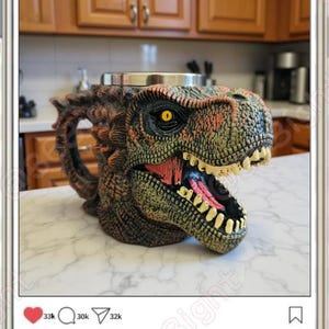 May include: A detailed, dinosaur-shaped mug with a textured, green, brown, and orange finish. The mug features a wide open mouth with visible teeth and a yellow eye. The mug is on a white surface, and the image is displayed on a phone screen.