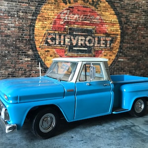 May include: A blue and white Chevrolet pickup truck model with a white cab and a blue bed. The truck is parked in front of a brick wall with a vintage Chevrolet logo painted on it.