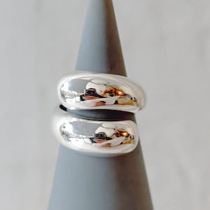 Könnte beinhalten: Ein silberner Ring mit zwei abgerundeten, überlappenden Bändern. Der Ring wird auf einem grauen, kegelförmigen Ständer präsentiert.