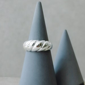 Peut inclure: Une bague argentée avec un motif torsadé est présentée sur un présentoir conique gris. La bague, au centre, met en valeur sa surface texturée. Un autre cône gris est en arrière-plan. Le fond est gris clair.