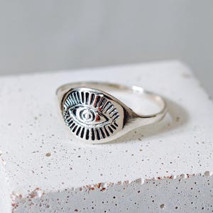 Può includere: Un anello d'argento con una faccia ovale raffigurante un occhio. Il design dell'occhio presenta linee dettagliate e una pupilla centrale. L'anello è esposto su una superficie bianca strutturata, evidenziando il design e l'artigianato.